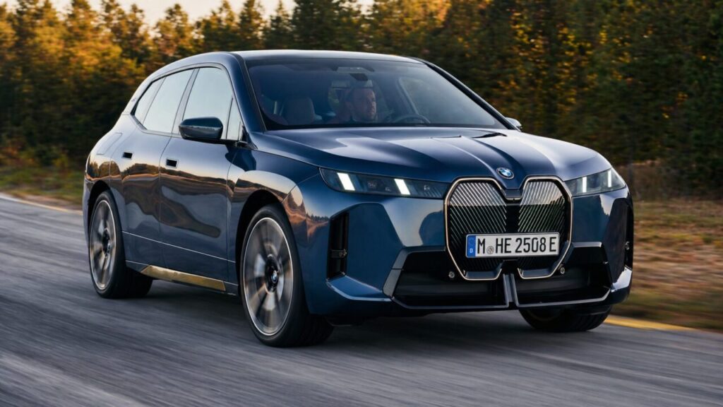Blue BMW iX electric SUV drives down a road at sunset in front of trees